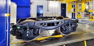 Railway bogie mounted on testing rig inside engineering laboratory, with blue and yellow machinery and industrial equipment visible.
